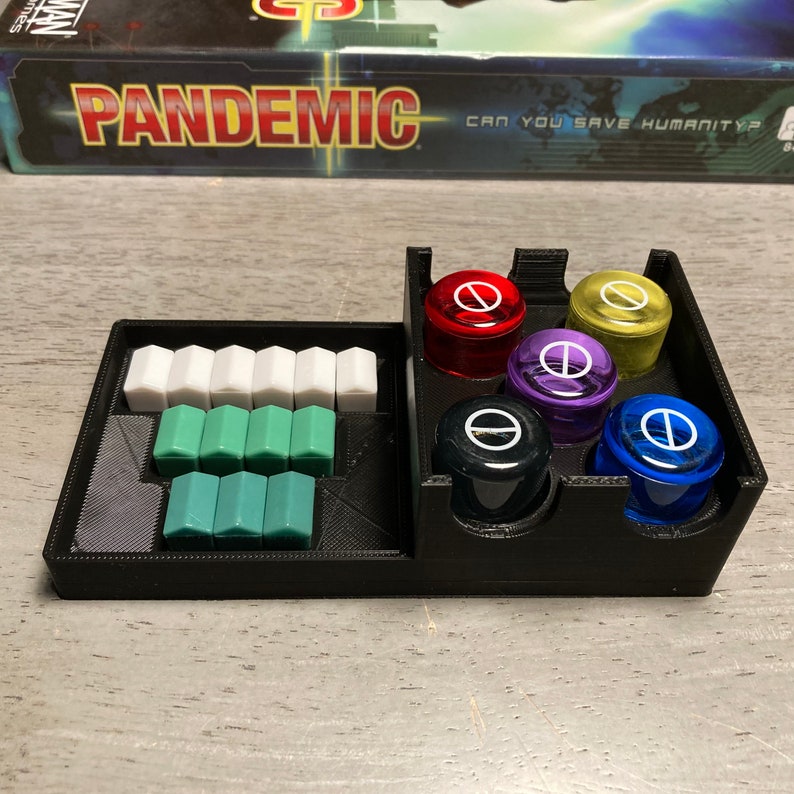 May include: A black plastic organizer for the board game Pandemic. The organizer has compartments for the game's colored cubes and player pieces. The game box is visible in the background with the text "Pandemic Can You Save Humanity?"