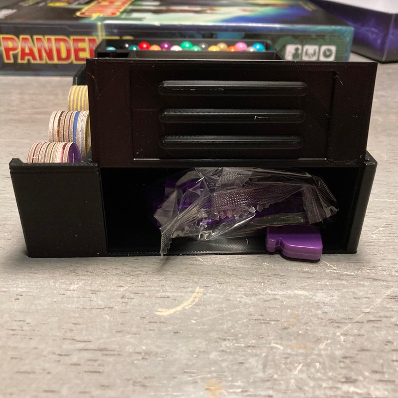 May include: A black plastic storage box with a compartment for small game pieces. The box has a clear plastic bag with purple game pieces inside.