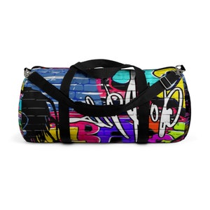 Könnte beinhalten: Eine zylindrische Sporttasche mit einem lebendigen Graffiti-Druck auf einem Ziegelsteinmuster. Das Design zeigt kräftige Spritzer in Blau, Pink, Gelb und Weiß mit schwarzen Akzenten. Sie verfügt über schwarze Nylongriffe und einen verstellbaren Schultergurt.