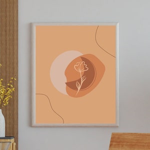 Può includere: Una stampa incorniciata con un design astratto che presenta un singolo fiore in uno schema di colori pesca, marrone e bianco. La stampa è appesa a un muro bianco sopra una sedia di legno.
