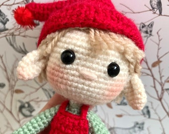 May include: A crocheted elf doll wearing a red hat, green and red overalls, and with black eyes. The doll is holding a small red pom-pom.