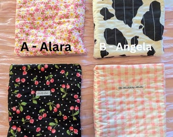 May include: Four patterned baby blankets are displayed. The blankets feature various designs: a pink daisy print, a black and white cow print, a black cherry print, and a pink gingham pattern. Each blanket has a name label: Alara, Angela, Nellie, and Lottie.