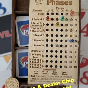 May include: A wooden game score tracker for Phases with a grid of holes for pegs, labeled with phase requirements and card values. The board includes a slot for card decks and a round wooden dealer chip. Text on the board reads Phases, Players, and Card Values.
