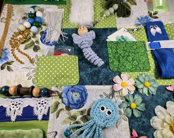 Puede incluir: Una manta de actividades sensoriales con un mosaico de telas florales y de lunares. Incluye un pulpo azul de ganchillo, una figura con cabeza de madera, varias cuentas, flecos blancos, encaje, bolsillos de tela y flores artificiales en la superficie.