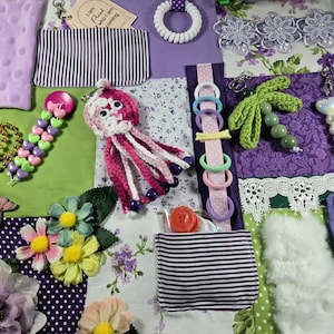 May include: A sensory activity mat featuring a patchwork of purple, green, and floral fabrics. It includes a crocheted pink octopus, a green crocheted palm tree, a white coil ring, fabric pockets, plastic beads, artificial flowers, and a tag with the text I am proud of who I am becoming.