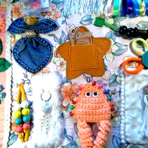 May include: A sensory activity mat featuring a floral fabric base with various attached elements including a denim angel, a felt star with a pocket containing a card that reads You are ENOUGH, a crochet monster, a white angel, beads, and ribbons.