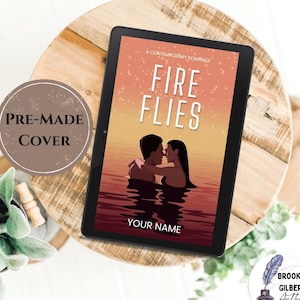 May include: A pre-made book cover mockup featuring a couple embracing in a lake at sunset with the title "Fire Flies" and the text "A Contemporary Romance". The cover is displayed on a wooden surface with a plant and a pre-made cover label.