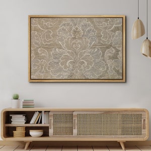 May include: A framed canvas print with a beige background and a white damask pattern. The print is hanging on a white wall above a wooden cabinet with woven rattan doors.