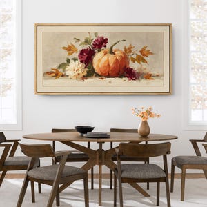 May include: A framed rectangular painting depicts a still life with a vibrant orange pumpkin, burgundy flowers, and autumn leaves. The artwork is set against a neutral background, with a gold-colored frame. The painting hangs above a wooden dining table.
