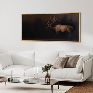 May include: A framed artwork featuring a majestic elk in a dark forest setting. The elk is depicted in profile, with large antlers and a light brown coat, set against a backdrop of shadowy trees. The frame is a warm, light brown color.