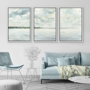 May include: A set of three framed abstract landscape paintings with a muted color palette of light blue, gray, and white. The paintings depict a seascape with a horizon line and clouds in the sky.