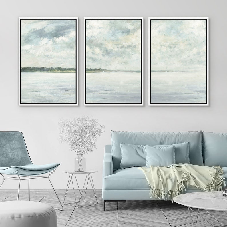 May include: A set of three framed abstract landscape paintings with a muted color palette of light blue, gray, and white. The paintings depict a seascape with a horizon line and clouds in the sky.