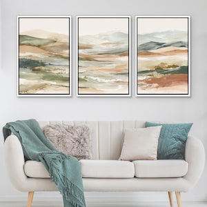 May include: Three framed landscape art prints. Each print features an abstract watercolor design with earthy tones of beige, brown, green, and blue. The artwork is displayed above a white sofa with decorative pillows and a teal throw blanket.