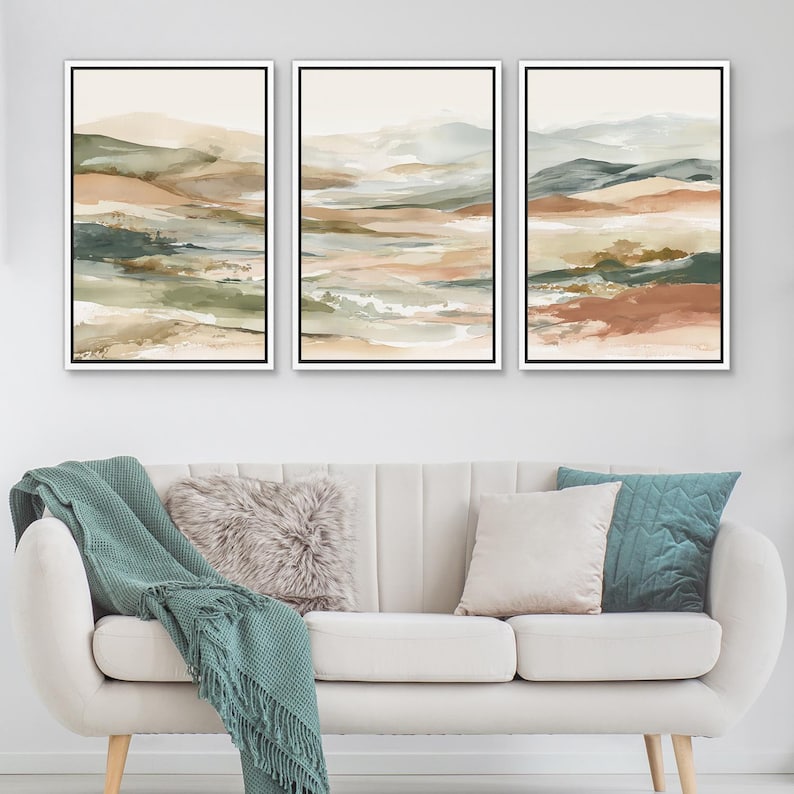 May include: Three framed landscape art prints. Each print features an abstract watercolor design with earthy tones of beige, brown, green, and blue. The artwork is displayed above a white sofa with decorative pillows and a teal throw blanket.