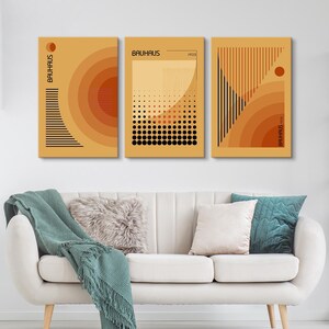 May include: A set of three rectangular wall art prints featuring Bauhaus-inspired geometric designs in shades of orange, yellow, and black. The prints display concentric circles, vertical lines, dot patterns, and the text Bauhaus 1923.