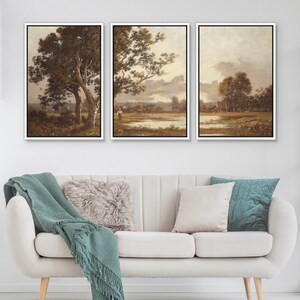 May include: Three framed landscape paintings with white borders. The artwork depicts trees, fields, and a body of water under a cloudy sky. The paintings are displayed above a white sofa with decorative pillows and a green throw blanket.