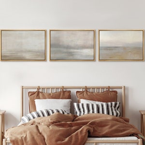 May include: Three framed abstract landscape paintings with muted tones of blue, gray, and brown. The paintings depict a seascape with a horizon line and a cloudy sky. The paintings are hung on a white wall above a wooden bed with brown bedding.