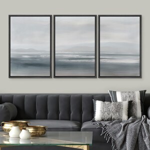 May include: A set of three framed canvas prints depicting a seascape with a hazy blue sky, a horizon line, and a body of water with white waves. The prints are displayed on a wall above a gray sofa with throw pillows and a blanket.
