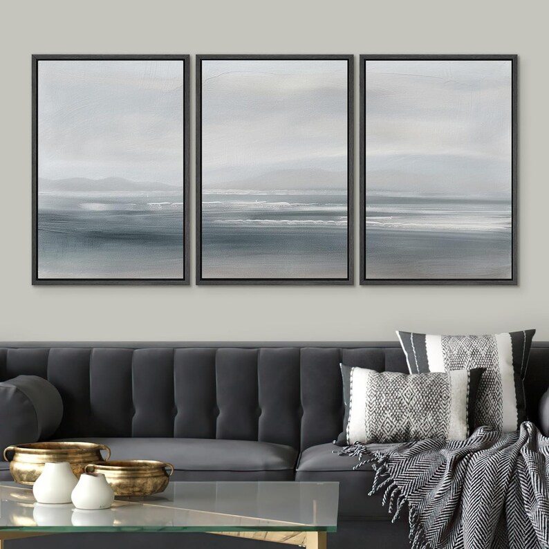 May include: A set of three framed canvas prints depicting a seascape with a hazy blue sky, a horizon line, and a body of water with white waves. The prints are displayed on a wall above a gray sofa with throw pillows and a blanket.