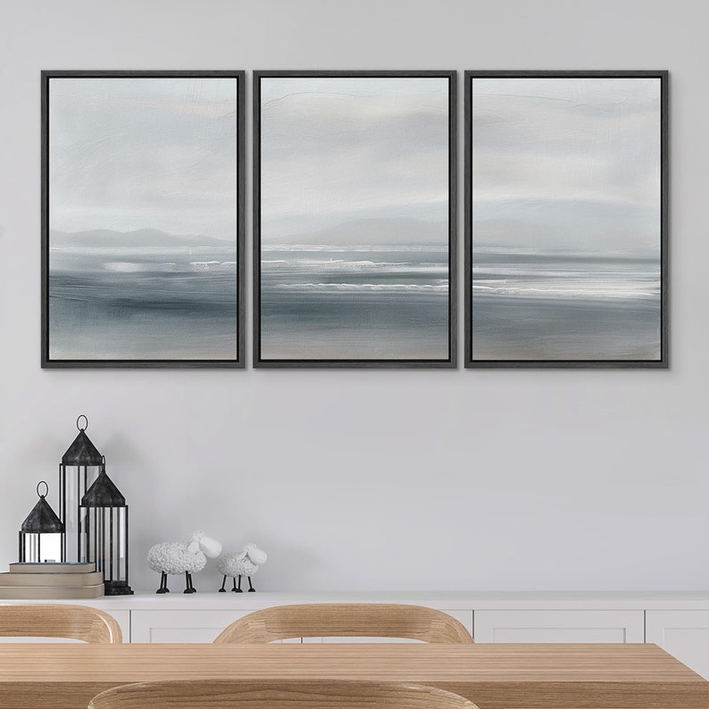May include: Three framed canvas prints with a seascape theme. The prints are in a muted blue and gray color scheme and depict a beach with waves and a distant horizon. The prints are arranged in a row on a white wall.