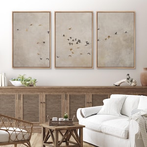 May include: Three framed prints of a flock of birds flying in formation against a light brown background. The prints are hung on a white wall above a wooden cabinet and a white couch.