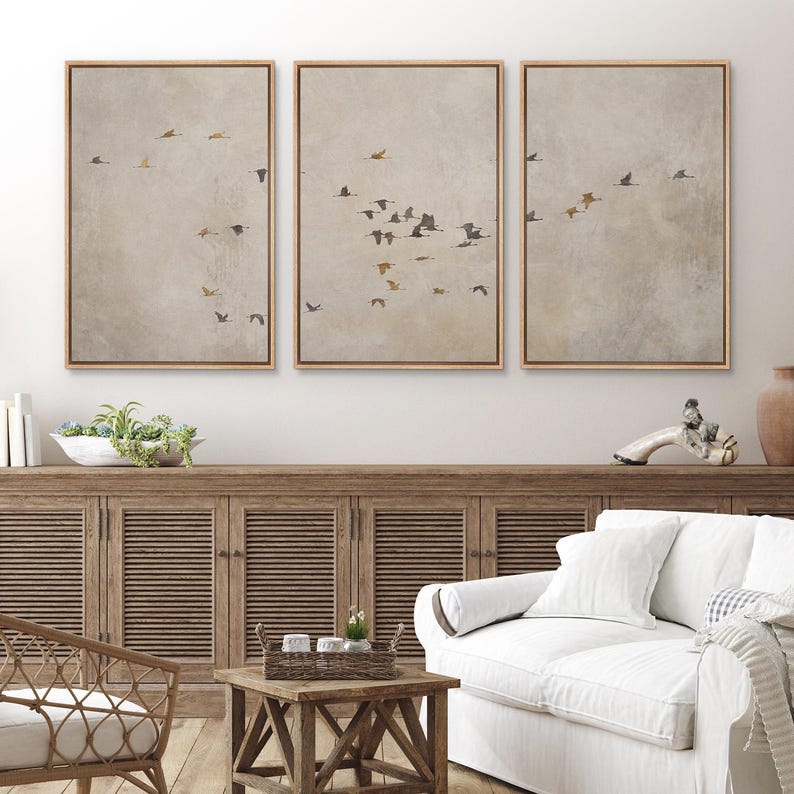 May include: Three framed prints of a flock of birds flying in formation against a light brown background. The prints are hung on a white wall above a wooden cabinet and a white couch.