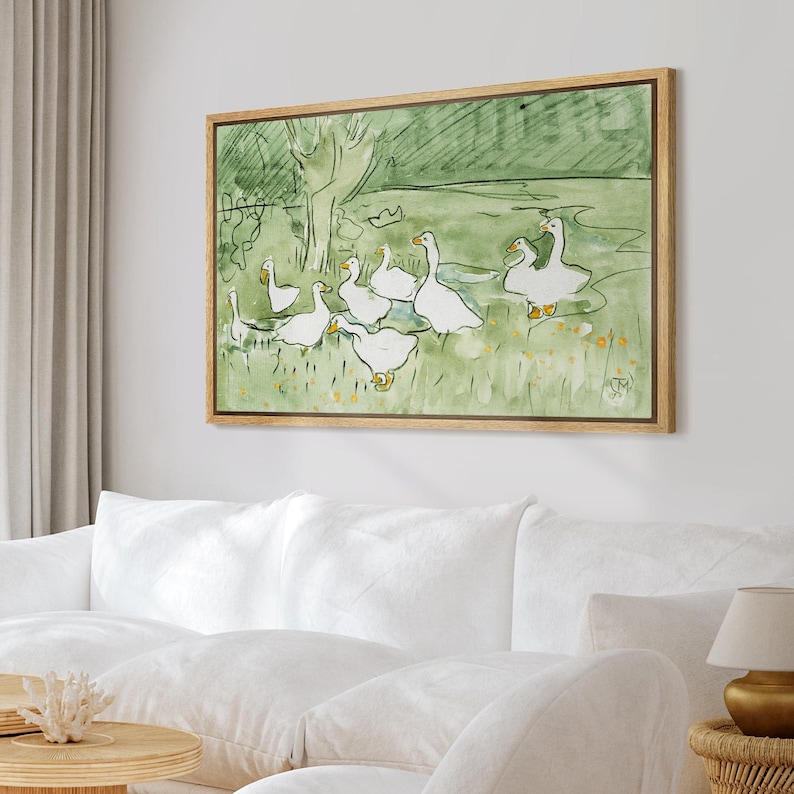 May include: A framed watercolor painting depicts a flock of white geese in a green field. The artwork features a light brown wooden frame. The geese are the focal point, with a simple, illustrative style. The background shows a tree and a grassy area.