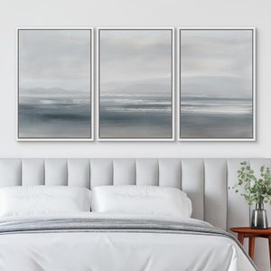 May include: A set of three framed canvas prints with a seascape theme. The prints are in shades of gray and white, depicting a horizon line with waves and a distant coastline. The prints are arranged in a row on a white wall above a white headboard.