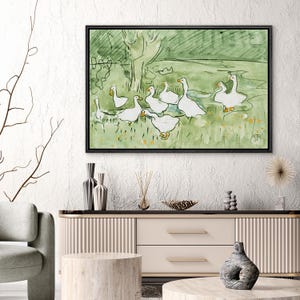 May include: A framed watercolor painting depicts a flock of white geese in a green field. The artwork features a simple, impressionistic style with a green and yellow color palette. The painting is displayed above a modern, neutral-toned cabinet.