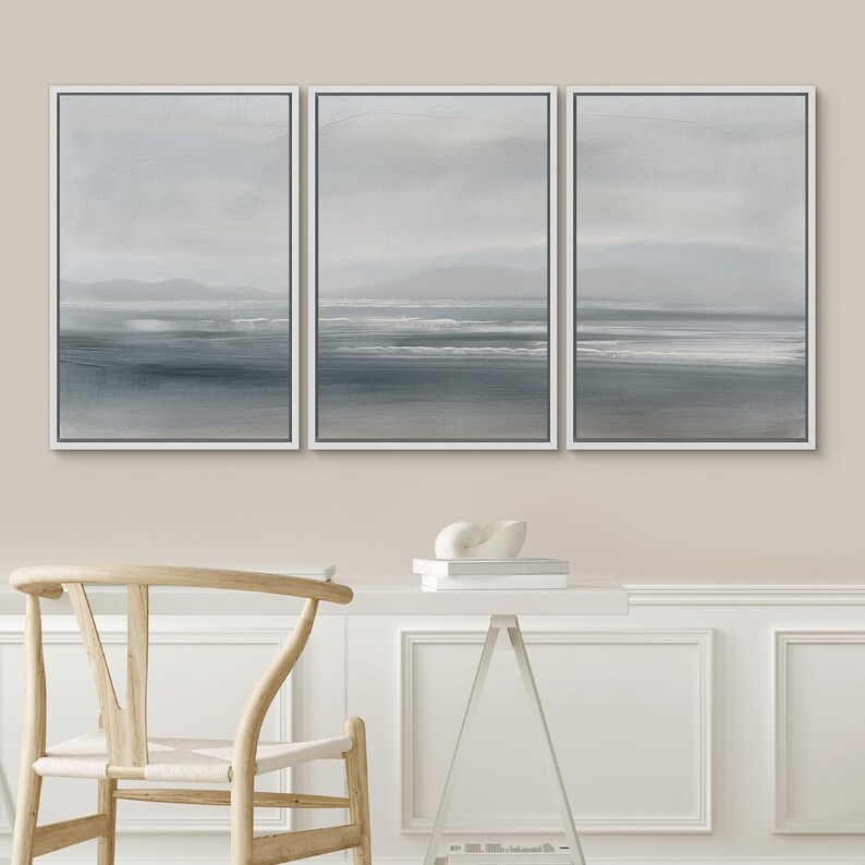May include: A set of three framed abstract landscape paintings. The paintings are in shades of gray and blue, with a subtle texture. The paintings are hung on a light beige wall.