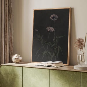 Puede incluir: Una impresi&oacute;n bot&aacute;nica enmarcada con dos flores moradas estilizadas con hojas verdes sobre un fondo oscuro. La obra de arte se exhibe en un mueble verde con un libro y un jarr&oacute;n decorativo.