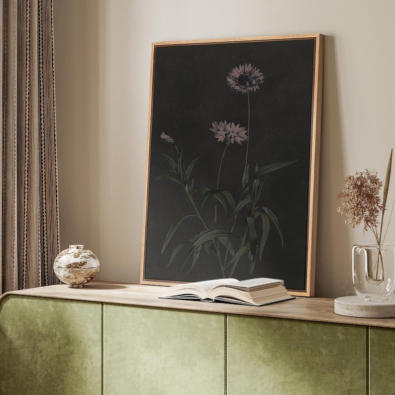 Puede incluir: Una impresi&oacute;n bot&aacute;nica enmarcada con dos flores moradas estilizadas con hojas verdes sobre un fondo oscuro. La obra de arte se exhibe en un mueble verde con un libro y un jarr&oacute;n decorativo.