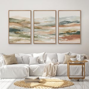 May include: Three framed landscape art prints. Each print features an abstract watercolor design with earthy tones of beige, green, and rust. The artwork is displayed above a white sofa with decorative pillows and a round woven rug.