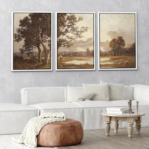 May include: Three framed landscape art prints. Each print features a sepia-toned pastoral scene with trees, fields, and a cloudy sky. The artwork is displayed above a white sofa, with a brown ottoman and small wooden table.