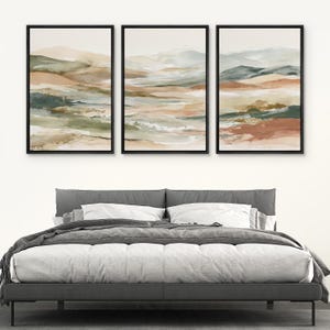 May include: Three framed abstract landscape paintings. Each features a watercolor style with muted tones of green, beige, and rust. The artwork is displayed above a gray bed frame with white bedding.