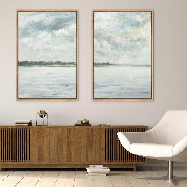 May include: Two framed abstract landscape paintings with a muted blue, green, and white color scheme. The paintings depict a calm body of water with a distant shoreline and a cloudy sky.