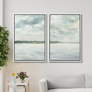 May include: A set of two framed landscape paintings depicting a serene seascape with a light blue sky, fluffy white clouds, and a calm body of water. The paintings are in a white frame with a black mat.