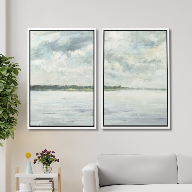 May include: A set of two framed landscape paintings depicting a serene seascape with a light blue sky, fluffy white clouds, and a calm body of water. The paintings are in a white frame with a black mat.