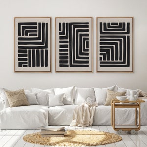 May include: Three black and white abstract geometric prints in wooden frames, hung on a white wall above a white couch with beige throw pillows and a beige throw blanket.