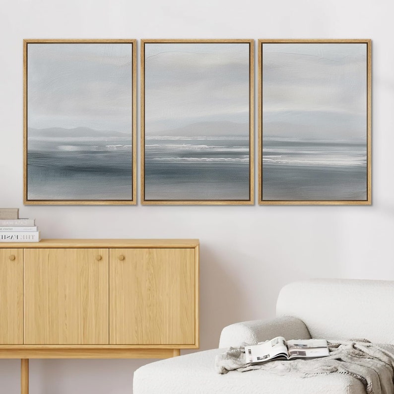 May include: Three framed canvas paintings with a minimalist seascape design. The paintings are in shades of gray and white, depicting a horizon line with a few waves and a distant shoreline. The paintings are hung on a white wall above a wooden cabinet.