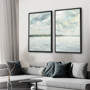 May include: Two framed abstract landscape paintings with a muted blue and green color scheme. The paintings depict a lake or ocean with a shoreline in the distance and a cloudy sky above.
