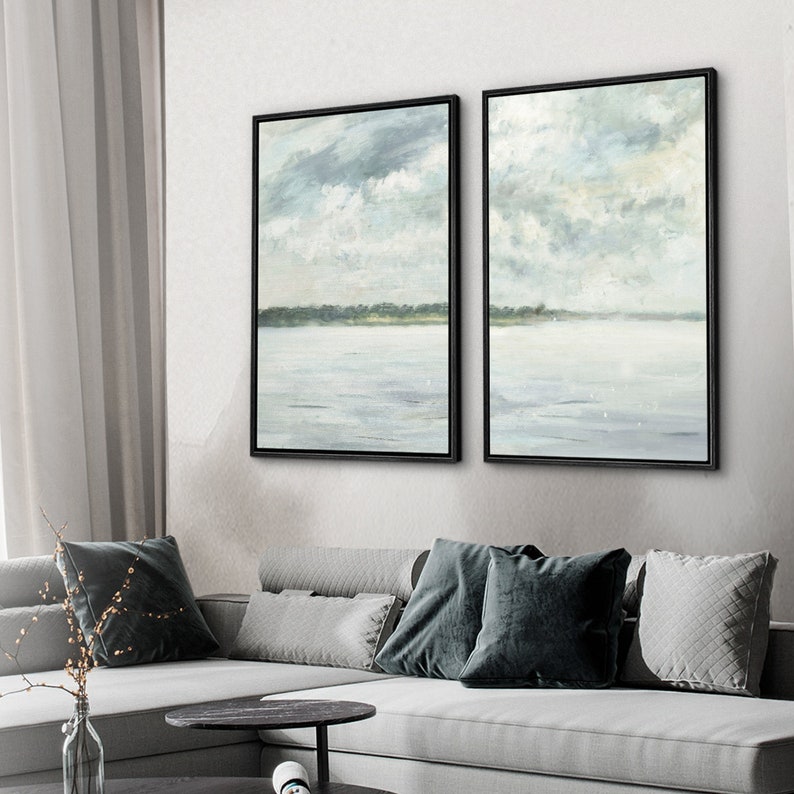 May include: Two framed abstract landscape paintings with a muted blue and green color scheme. The paintings depict a lake or ocean with a shoreline in the distance and a cloudy sky above.