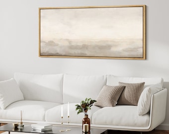 Pode incluir: Uma pintura abstrata horizontal em tons suaves de bege e marrom, emoldurada em uma moldura de madeira clara, pendurada acima de um sofá branco com almofadas.
