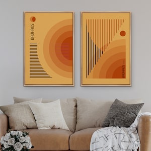 May include: Two framed rectangular wall art prints featuring geometric designs in shades of yellow and orange. The left print displays the text BAUHAUS with concentric circles and horizontal lines. The right print shows the text BAUHAUS 1923.