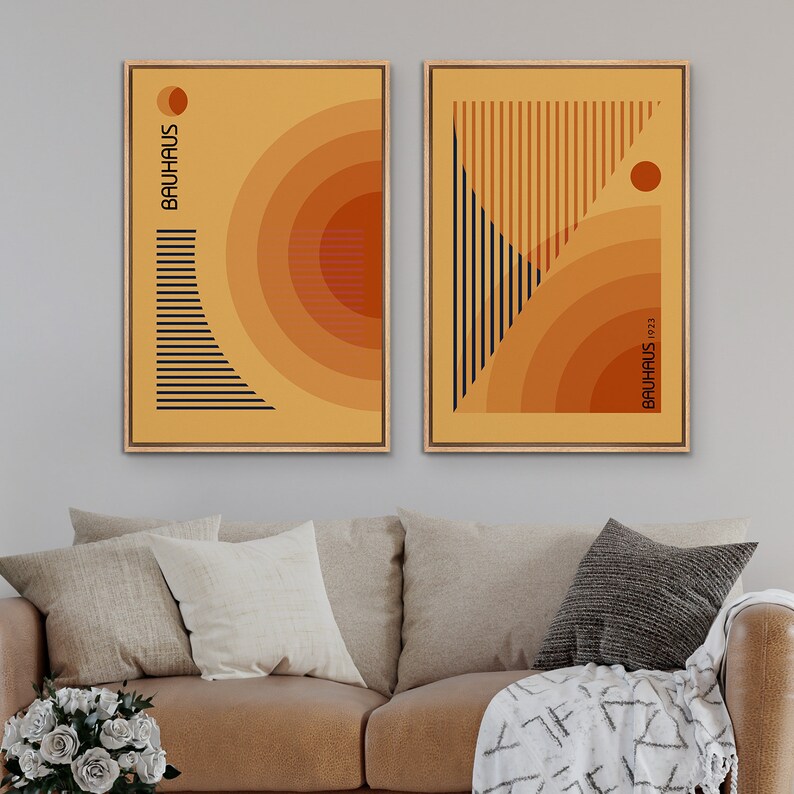 May include: Two framed rectangular wall art prints featuring geometric designs in shades of yellow and orange. The left print displays the text BAUHAUS with concentric circles and horizontal lines. The right print shows the text BAUHAUS 1923.