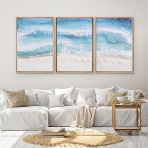 May include: Three framed wall art pieces depicting an aerial view of a beach with turquoise water and white waves. The artwork is framed in light brown wood and hangs above a white sofa with decorative pillows.