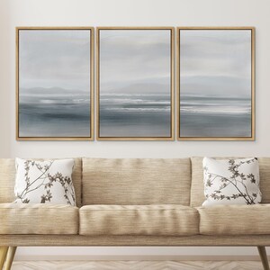 May include: Three framed canvas prints of a seascape with a light blue and gray color scheme. The prints are arranged in a row on a white wall above a beige couch.