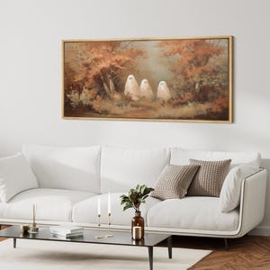 May include: A framed canvas art print featuring three white ghost figures in a forest setting with autumn foliage. The artwork has a warm color palette with shades of brown, orange, and green, and is displayed above a white sofa.