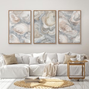 May include: Three framed abstract art prints with a watercolor design in neutral tones of beige, gray, and white. The artwork is displayed above a white sofa with decorative pillows, a round woven rug, and a wooden side table.