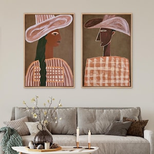 May include: Two framed rectangular wall art prints featuring stylized portraits of individuals wearing wide-brimmed hats. The left figure has a long dark green braid, while the right figure has a sharp profile. Both wear patterned tops in earth tones.